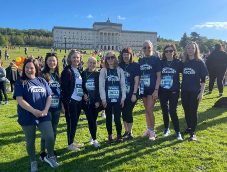 A group of 9 women stand in front of the Stormont Parliament Buildings in their ASCERT t-shirts at the beginning of the Belfast Marathon.