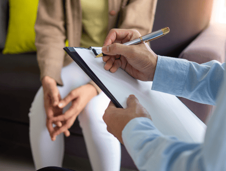 A counsellor speaking to a client. We do not see their faces. The counsellor writes on a clipboard while the client has their hands interlaced.