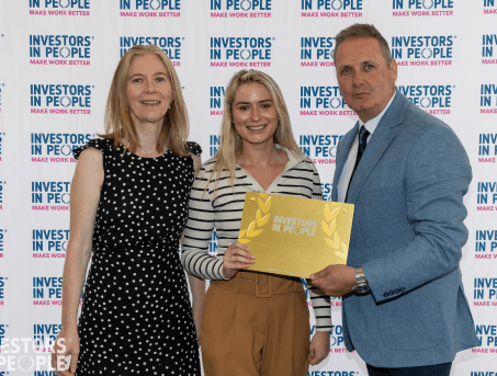 Nuala and Ella from ASCERT's HR Team stand with a man presenting them with an Investors In People Gold Award.