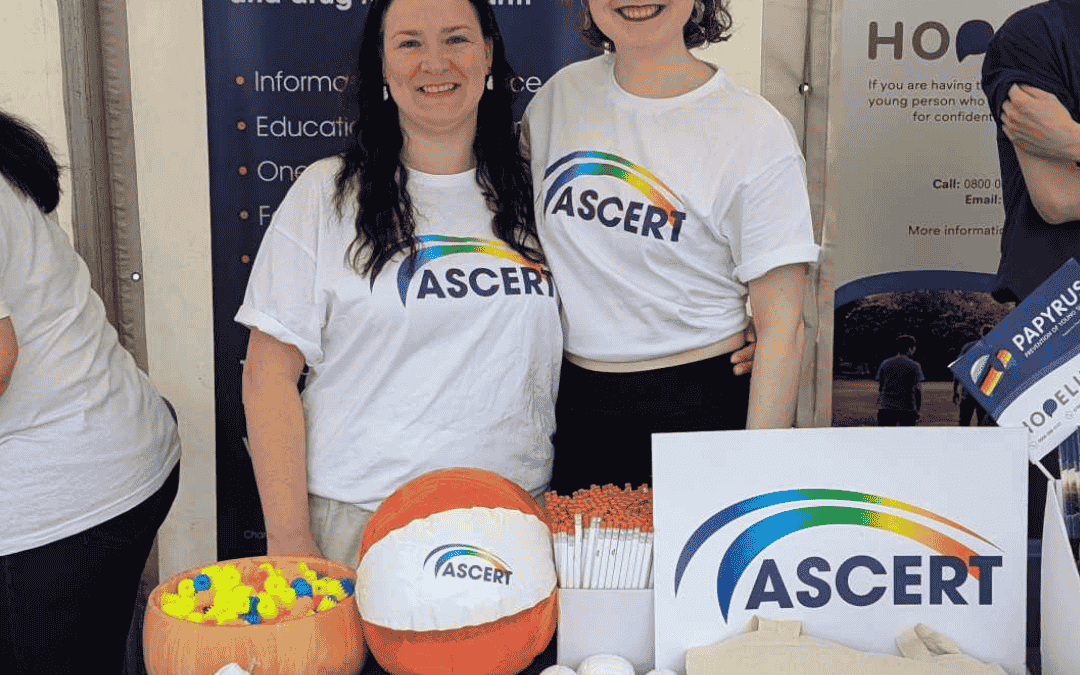 Two women who are ASCERT staff members smile for a photo. They are wearing white t-shits with a rainbow coloured ASCERT logo. They stand in front of an ASCERT roller banner and an ASCERT merchandise table.