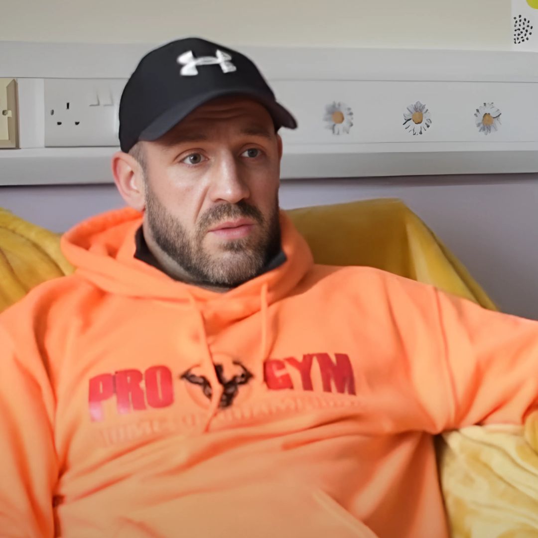 A man sits on a yellow chair. He wears a black baseball cap and a orange huddie.