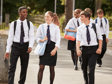 Three teenagers in school uniforms, two boys and a girl, walking on a footpath. They wear a white shirt with a black striped tie and back trousers. They all wear schoolbags.
